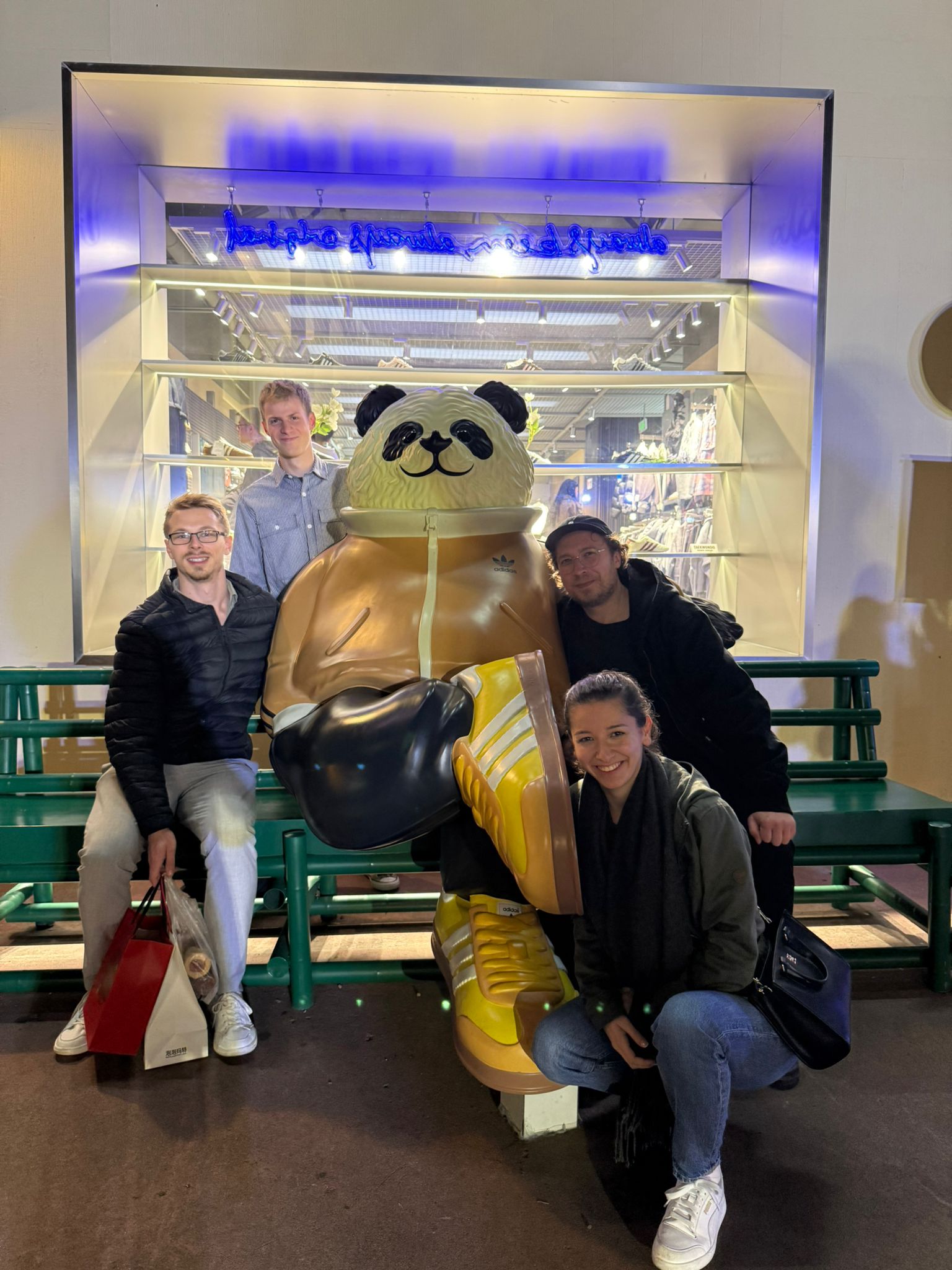 A foto of Luiz, Bastian, Max (males) and Sümeyye (female) with a statue of a panda bear dressed in adiddas clothes and shoes