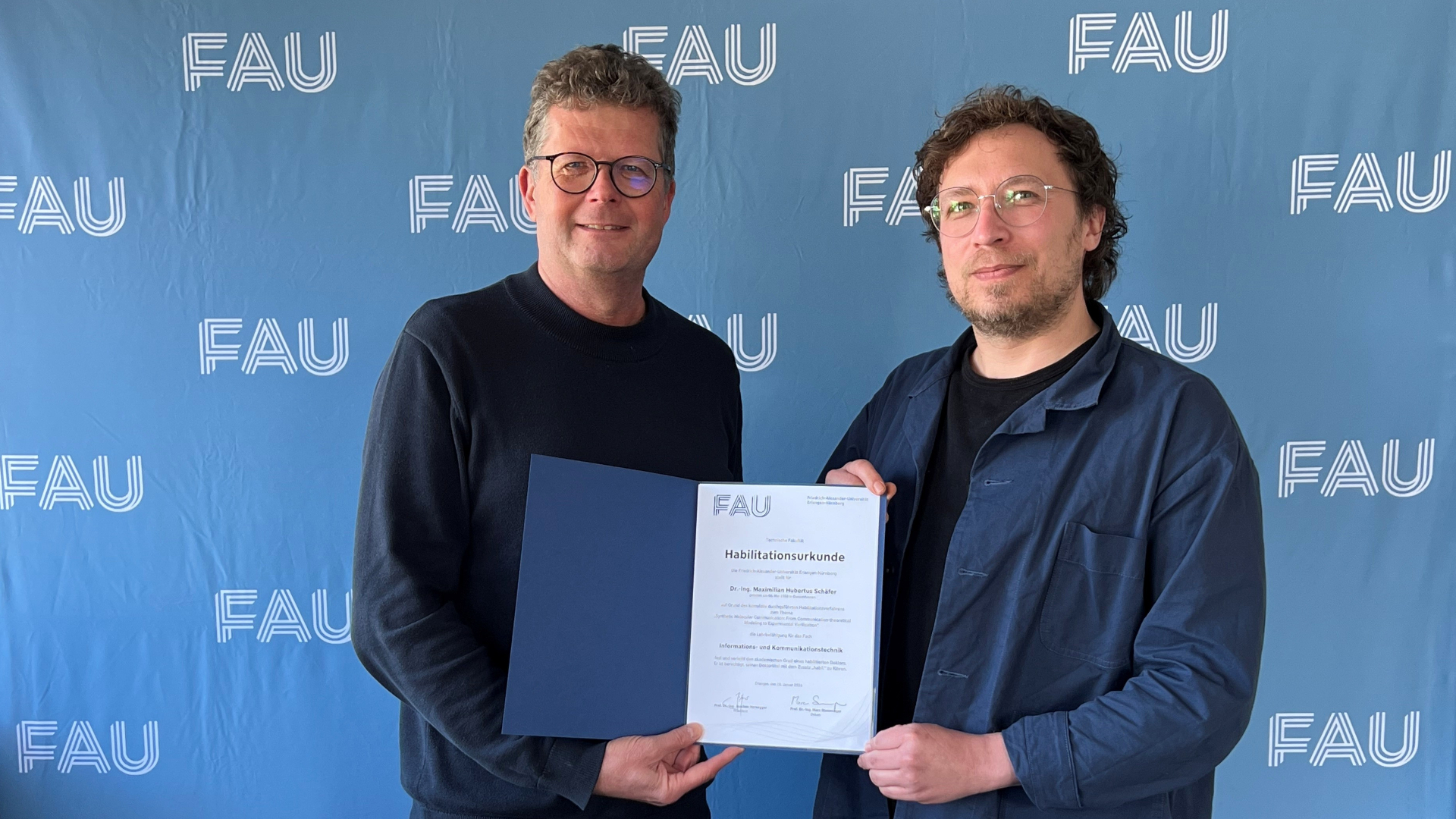 Dr.-Ing. habil Maximilian Schäfer (right, male) with his habilitation certificate and The dean of the Faculty of Engineering, Prof. Dr.-Ing. Marc Stamminger (left, male) in front of the blue FAU background