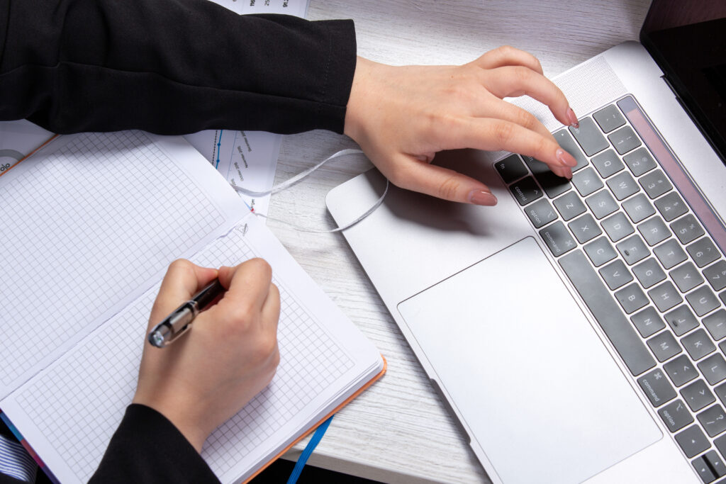 Hands of a person - one typing on a laptopt and the other making notes in a notebook
